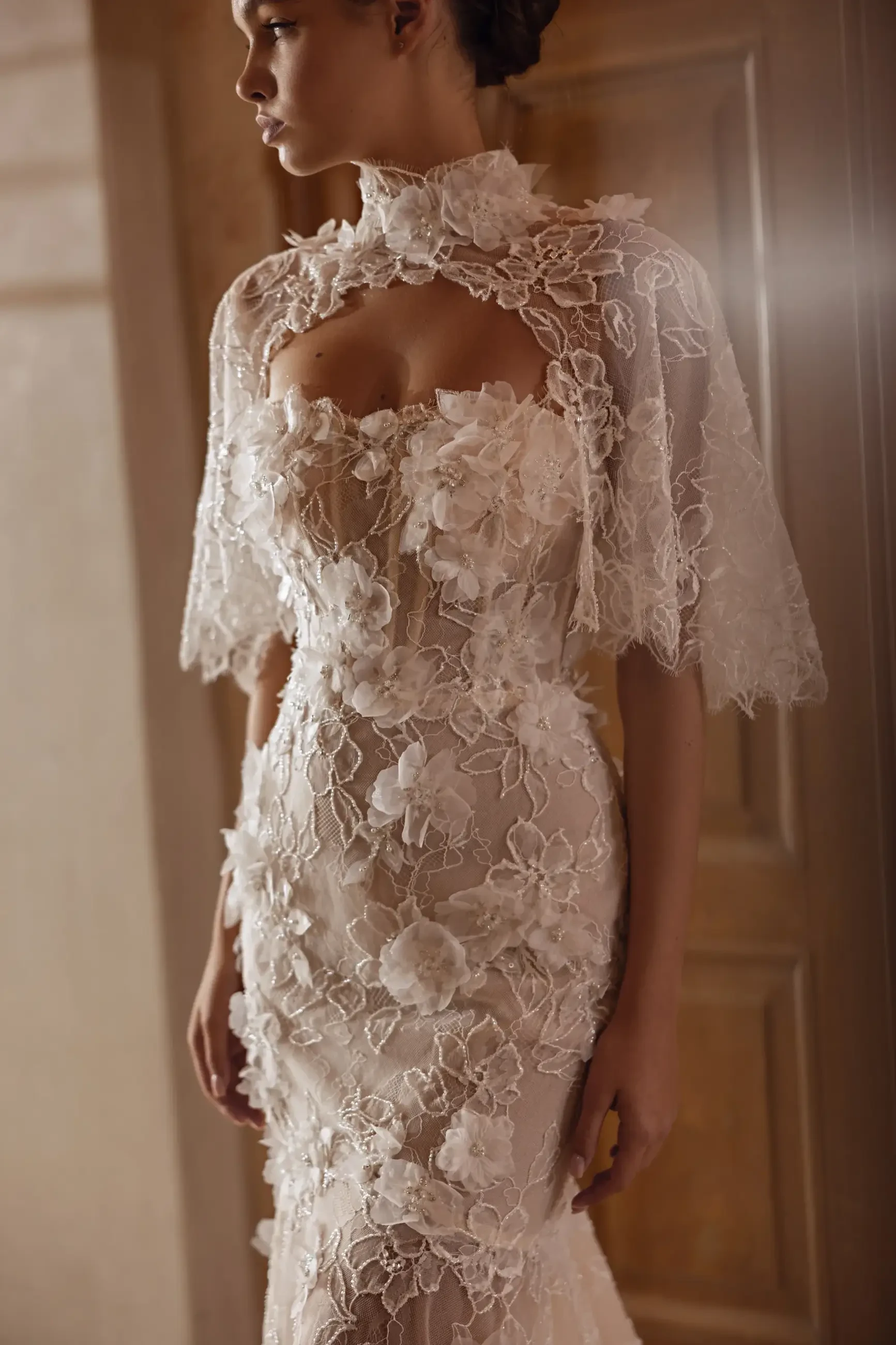 A woman wearing an elegant, lace-embroidered gown with floral patterns, standing near a wooden door. The mood is romantic and sophisticated.