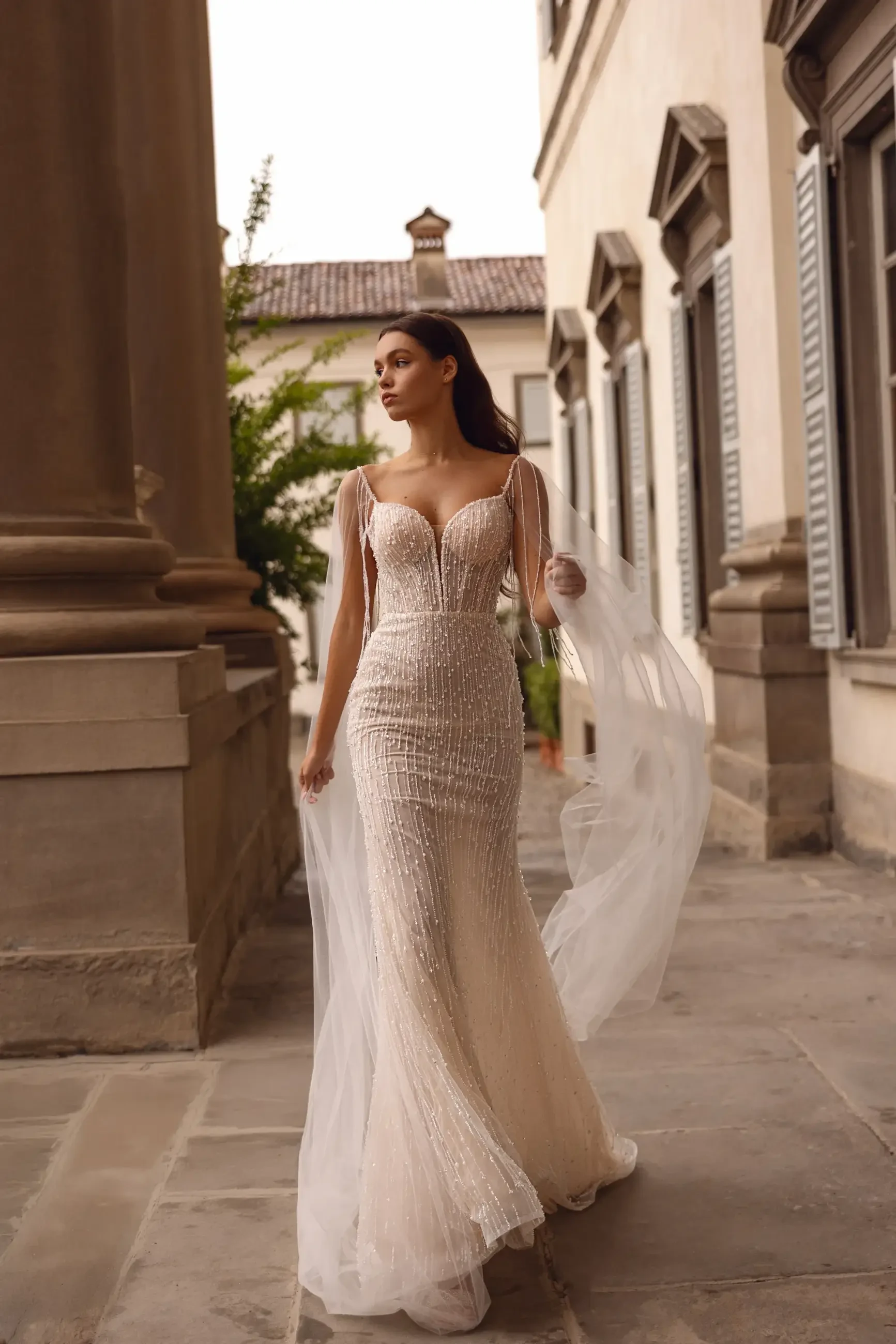 A woman in an elegant, form-fitting wedding dress with lace details walks gracefully through a courtyard surrounded by historic architecture.