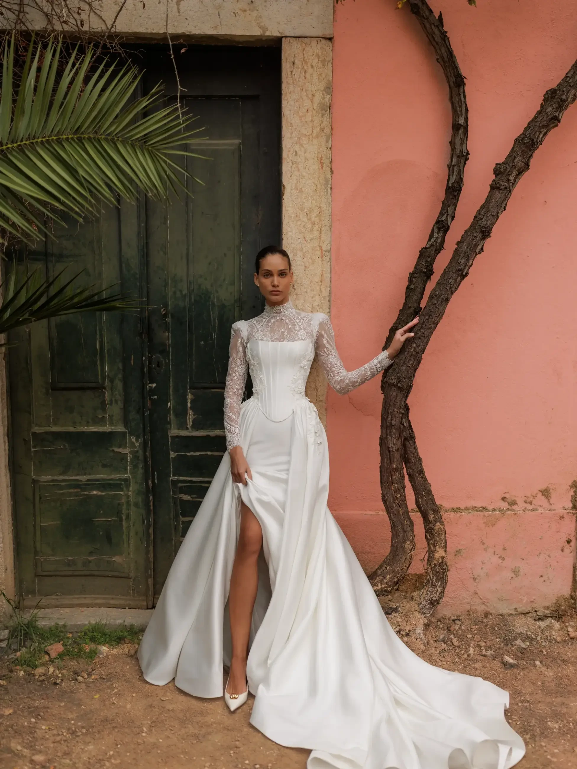 A woman in an elegant wedding gown with a lace bodice and long train stands confidently by a rustic green door and pink wall. A palm frond adds texture.