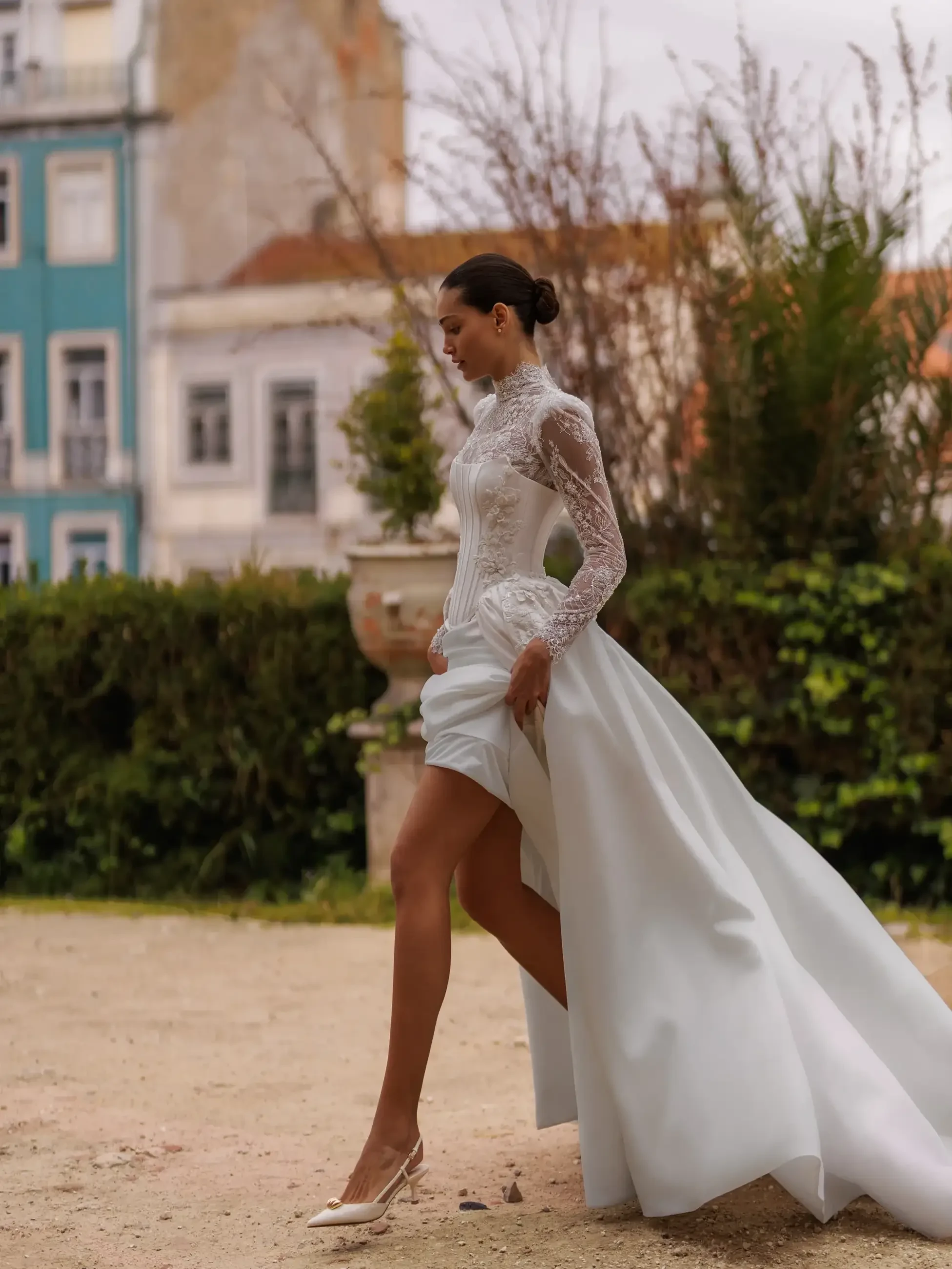 A model walking in a stylish wedding dress with a high-low hemline and lace details, set in a picturesque outdoor location with greenery and historic buildings in the background.