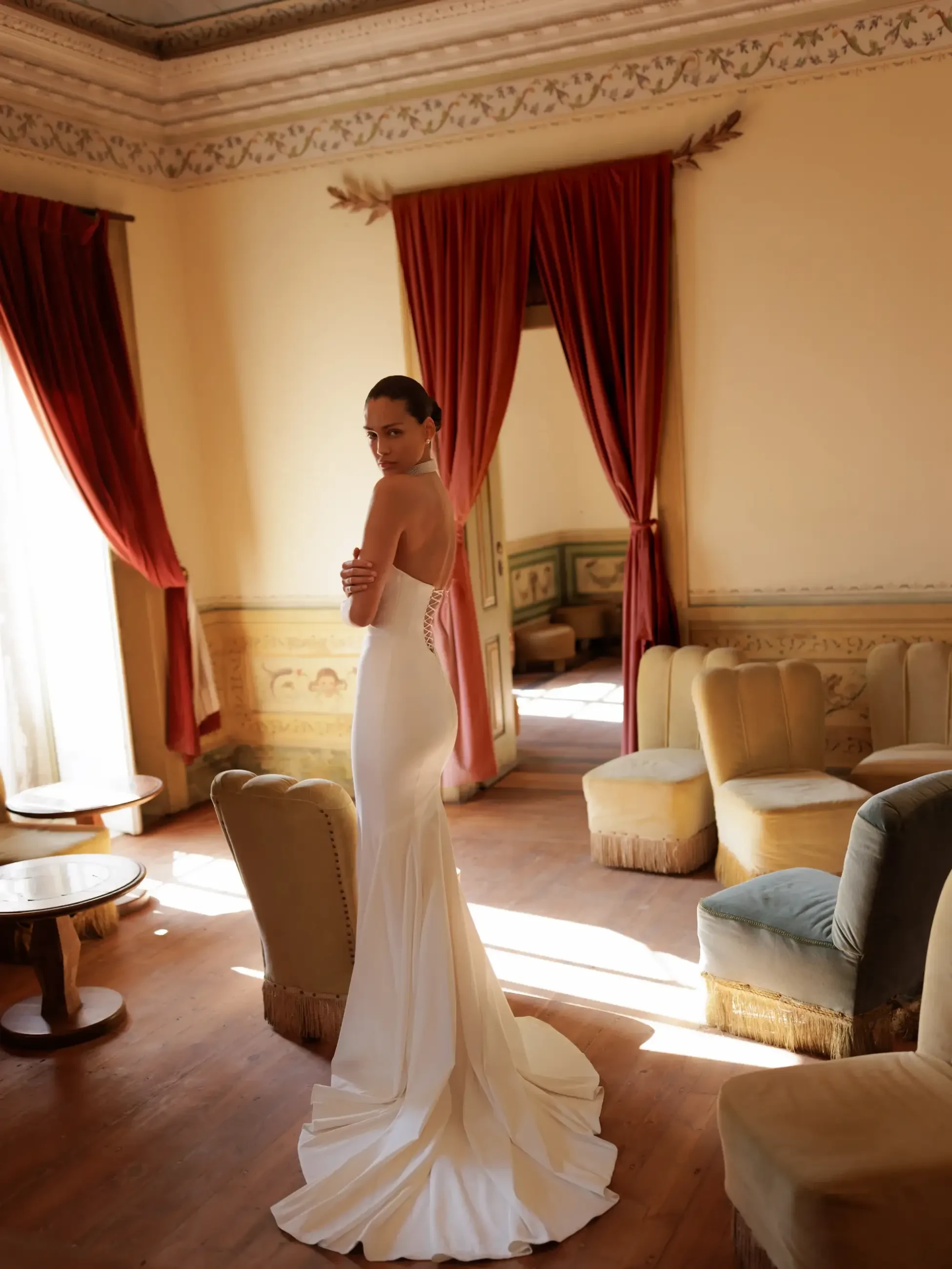 A woman in a white dress stands in an elegant room with curtains and drapery, looking over her shoulder. The room has chairs and tables arranged around her.