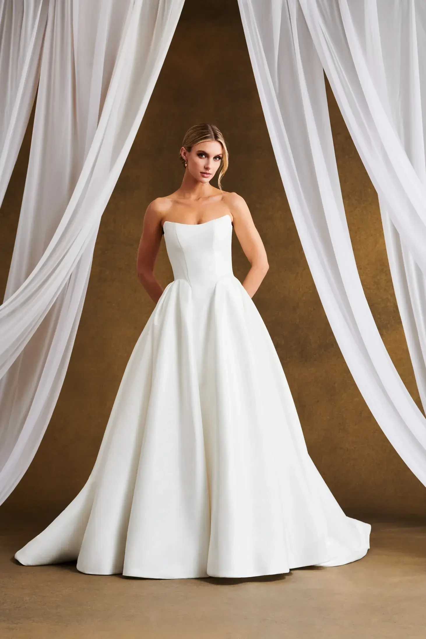 A model wearing a strapless white wedding gown, standing gracefully in front of a backdrop with draped fabric.