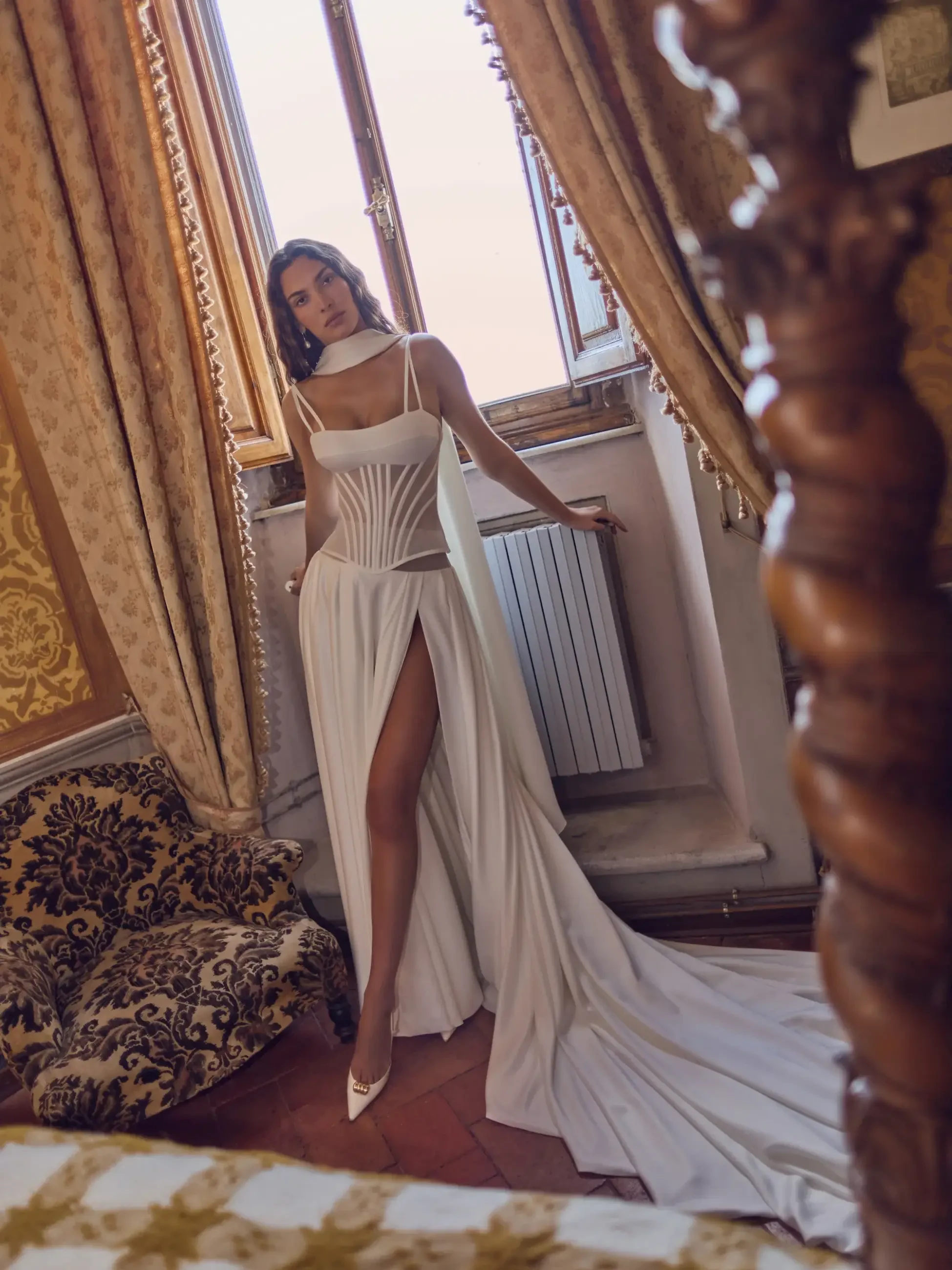 Model is wearing a flowing satin A-line gown with a sheer corset bodice, delicate straps, a thigh-high slit, and a dramatic detachable back shawl.