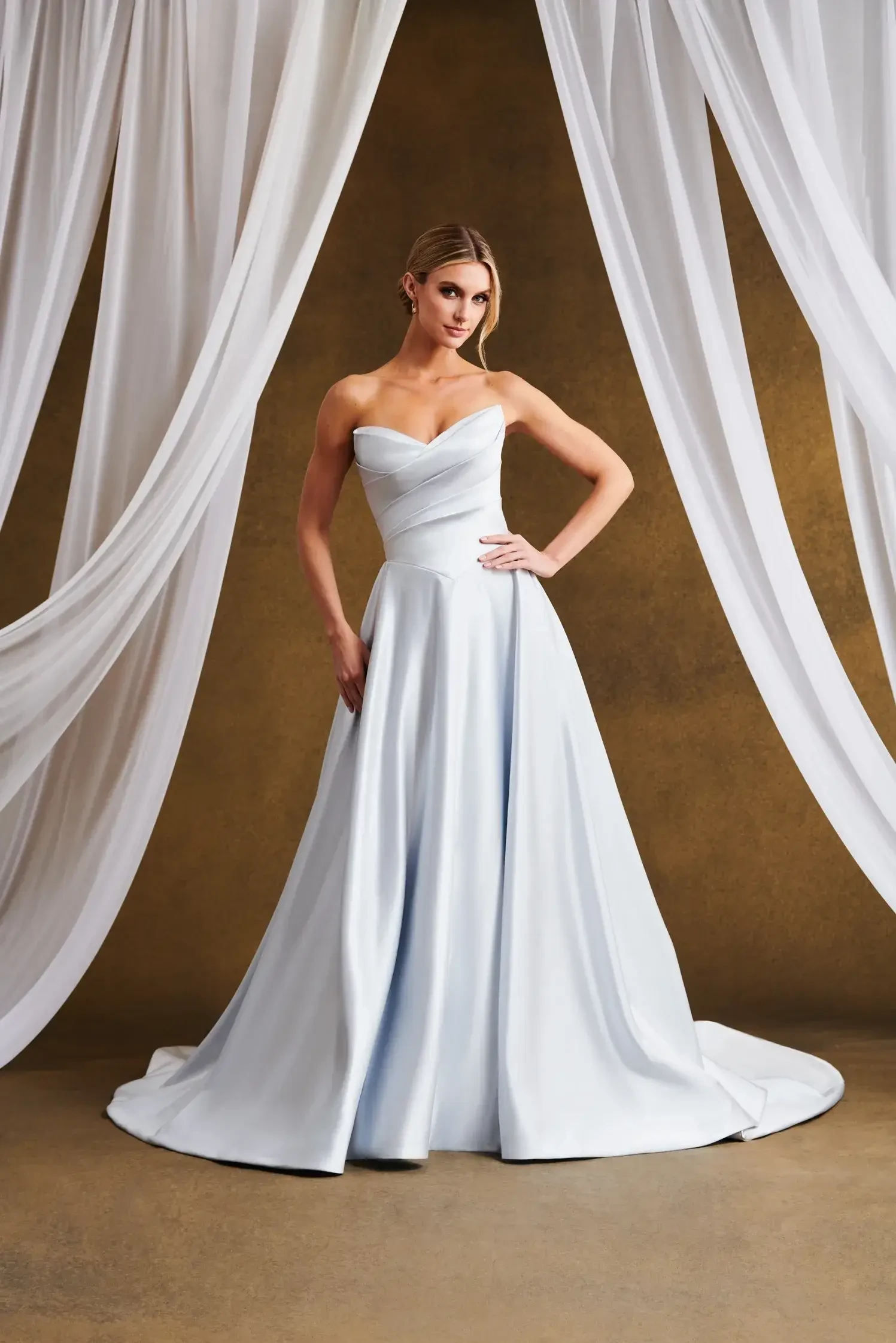 A woman in an elegant light blue strapless gown stands confidently against a brown backdrop, flanked by flowing white drapes, evoking a serene and refined atmosphere.