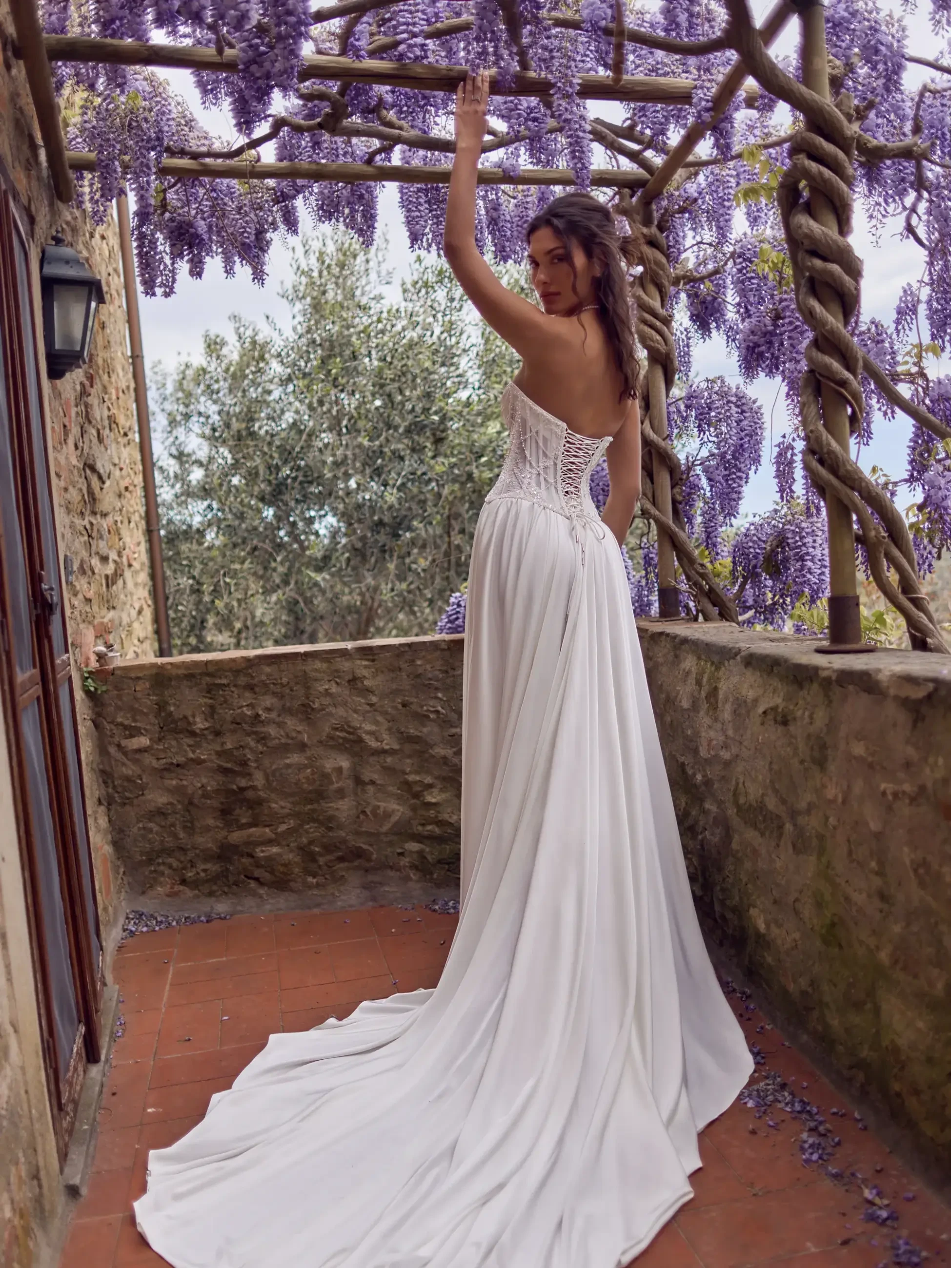 Model is wearing a romantic gown with a sheer lace-up corset and light silk skirt, styled with a removable lace overlay for two distinct bridal looks.