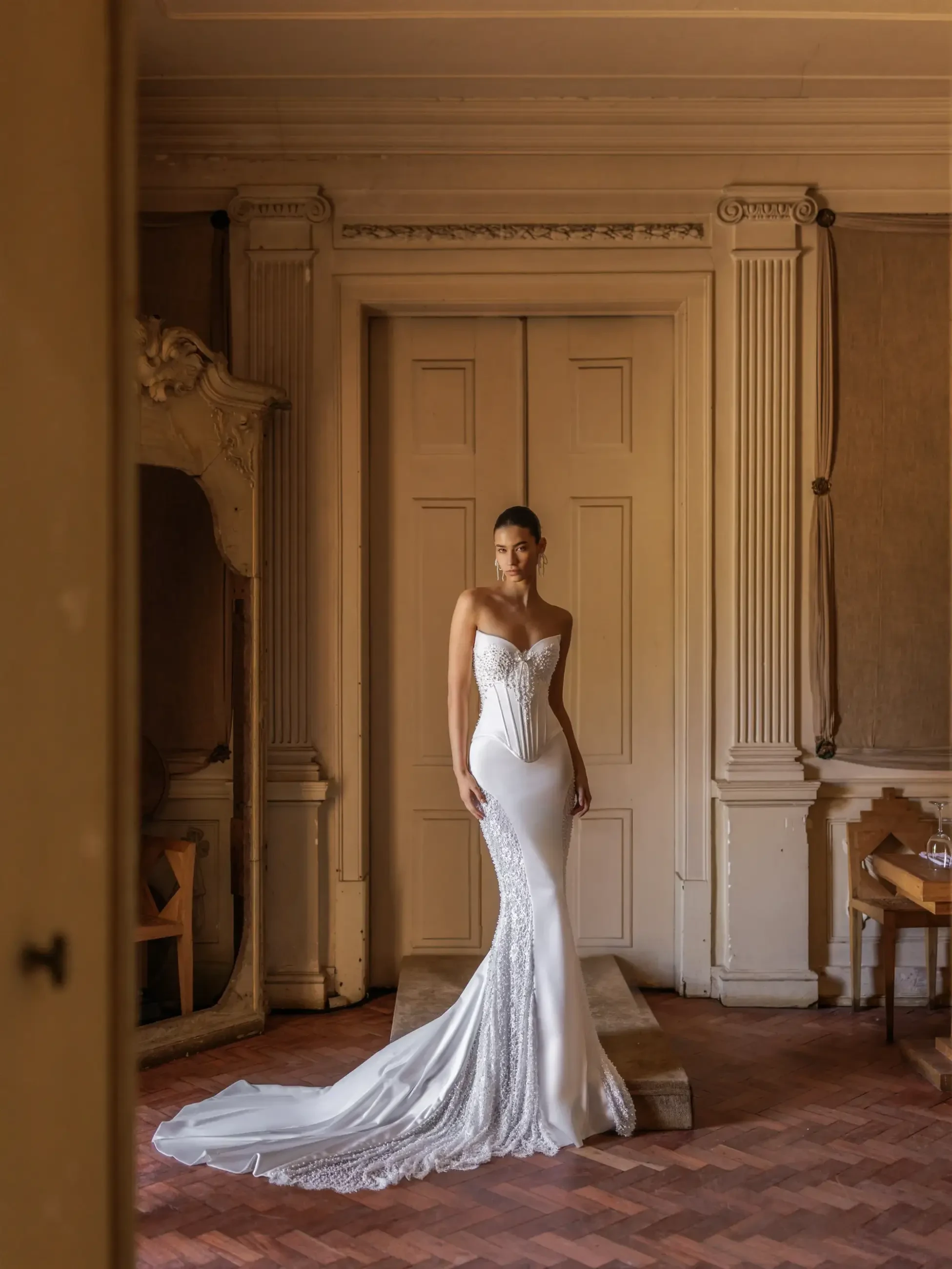 A bride in a strapless, form-fitting white gown with lace details stands in an elegant room with ornate pillars, exuding a graceful and serene ambiance.